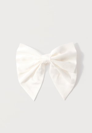 Krémová tkaninová stuha do vlasov s malým strieborným príveskom v tvare koruny na uzle, zobrazená na svetlošedom pozadí.