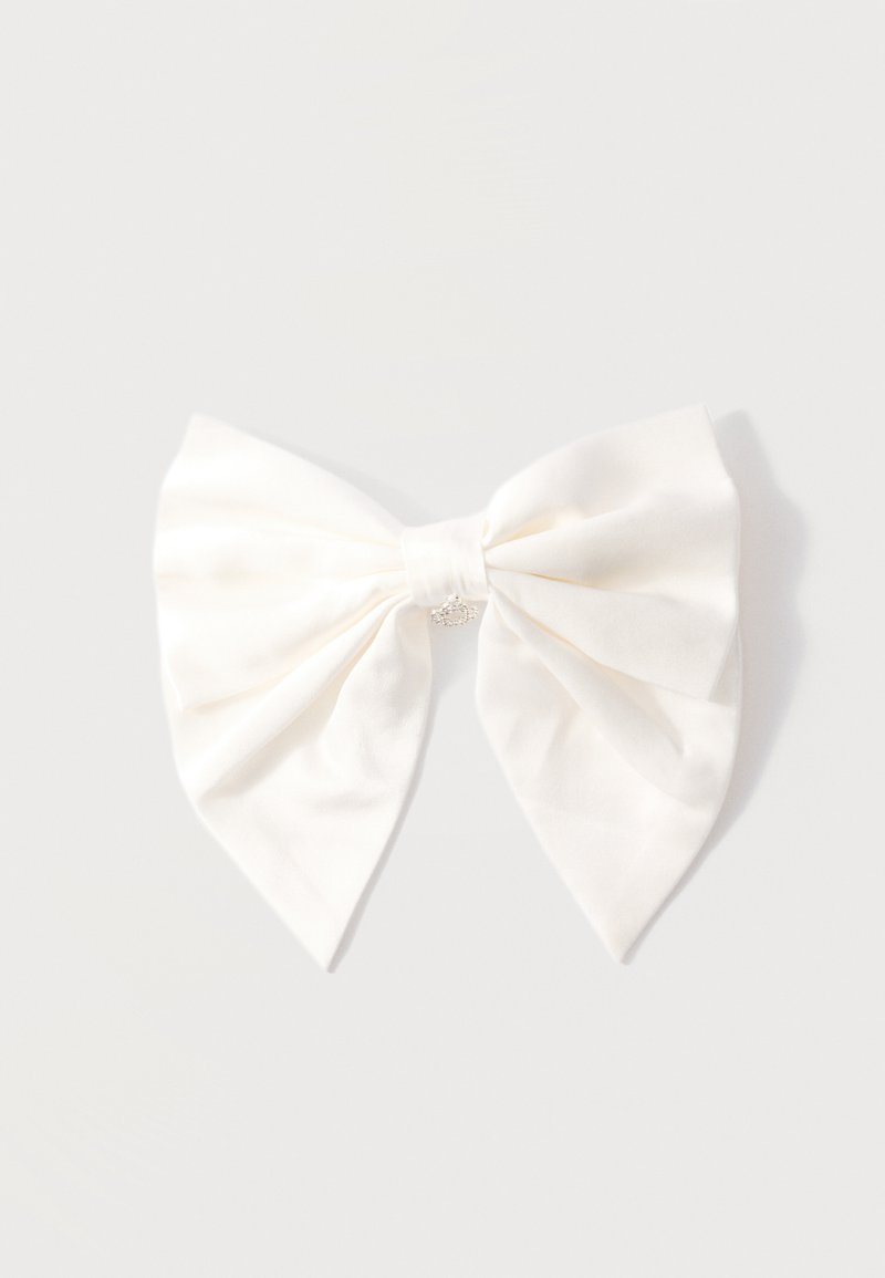 Nœud à cheveux en tissu crème avec un petit charme en forme de couronne argentée au nœud, présenté sur un fond gris clair.