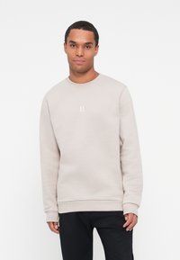 Sudadera gris claro hecha de tejido suave, con cuello redondo, puños acanalados y un pequeño logo blanco centrado en el pecho.