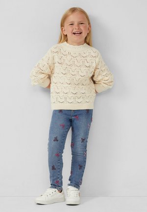 Pull en tricot crème avec un motif texturé, associé à un jean bleu orné de dessins de nœuds brodés roses. Complété par des baskets blanches.