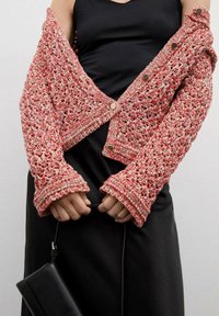 Gehaakte cardigan in rood en crème met grote manchetten en knoopsluiting, gedragen over een zwarte top. Zwarte handtas in de handen vastgehouden.