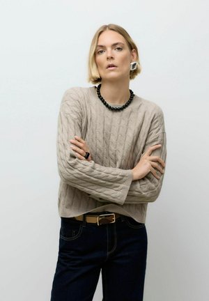 Beiger Kabelstrickpullover mit langen Ärmeln, kombiniert mit dunklen Jeans und einer schwarzen Perlenkette. Einfaches, strukturiertes Design, ohne Muster.