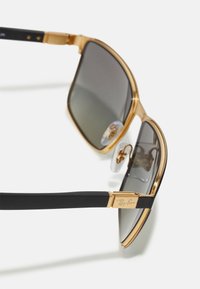 Ochelari de soare aurii cu brațe negre, având lentile gri, o formă rectangulară subțire și pernițe transparente pentru nas. Logo-ul Ray-Ban pe tâmplă.