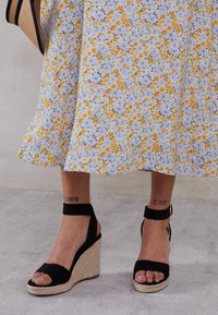 Sandalias de cuña negras con suela de esparto texturizada y tiras en el tobillo, combinadas con una falda de estampado floral con flores amarillas y azules.