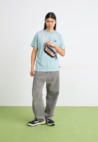 Light blue cotton t-shirt with a logo, gray corduroy pants, and a multicolored crossbody bag. Black sneakers complete the outfit.