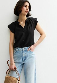 Blusa negra de manga corta con cierre de botones, con un cuello fruncido, combinada con jeans de cintura alta de color azul claro y un bolso de mimbre.