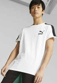 Camiseta blanca de manga corta con detalles en negro en los hombros y el logo de Puma. Fabricada en algodón, cuenta con un ajuste relajado y un diseño de cuello redondo.
