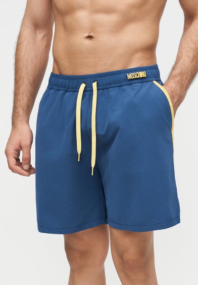 Męski tors w niebieskich kąpielówkach Moschino z żółtymi sznureczkami i bocznym paskiem, jedna ręka w kieszeni, na tle jednolitego jasnego tła.