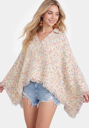 Giovane donna che indossa un poncho lavorato a maglia multicolore con frange sopra pantaloncini di jeans strappati e orecchini arancioni a forma di fiore, sorridendo e guardando di lato.