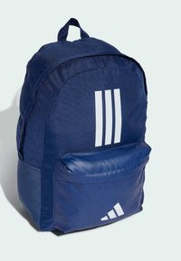 Granatowy plecak z tkaniny, okrągły kształt i białe akcenty, w tym trzy pionowe paski i logo na przedniej kieszeni.