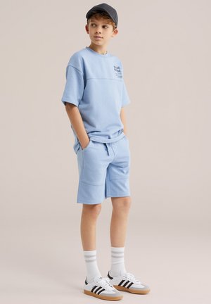 Ragazzo che indossa pantaloncini e maglietta coordinati azzurri, cappello grigio, calzini bianchi alti e scarpe da ginnastica bianche con strisce nere, in piedi con le mani nelle tasche.