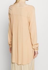 Chemise à manches longues couleur pêche en tissu léger, présentant une coupe ample, des poignets boutonnés, des plis au dos et un ourlet arrondi.