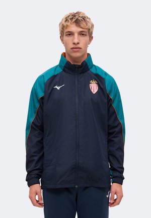 Un jeune homme porte une veste de sport zippée de l’AS Monaco, bleu marine et bleu sarcelle, sur un fond clair uni.