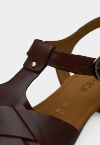 Καφέ δερμάτινο σανδάλι με σχέδιο T-λουρί, στρογγυλή αγκράφα και λείο επεξεργασμένο πέλμα με χρυσές εντυπωμένες λεπτομέρειες μεγέθους.