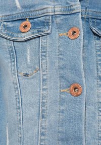 Veste en denim bleu clair avec boutons en cuivre et coutures dorées, dotée de deux poches poitrine et de détails usés sur le tissu.