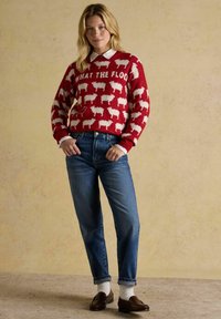 Frau in einem roten Pullover mit weißen Schafen und dem Text "WHAT THE FLOCK", blauen Jeans, weißen Socken und braunen Loafers, die gegen eine beige Wand steht.
