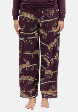 Personne portant un pantalon large violet foncé avec un motif de léopards se prélassant sur des branches d'arbres, debout pieds nus.