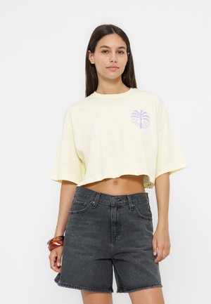 Kvinde iført en lysegul cropped T-shirt med lilla palme grafik og højtaljede mørkegrå denimbukser, stående mod hvid baggrund.