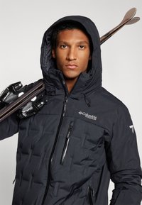 Crna izolirana jakna s kapuljačom, s teksturiranim uzorkom, zatvorenim džepovima i Columbia logom na prsima. Skije su prebačene preko jednog ramena.