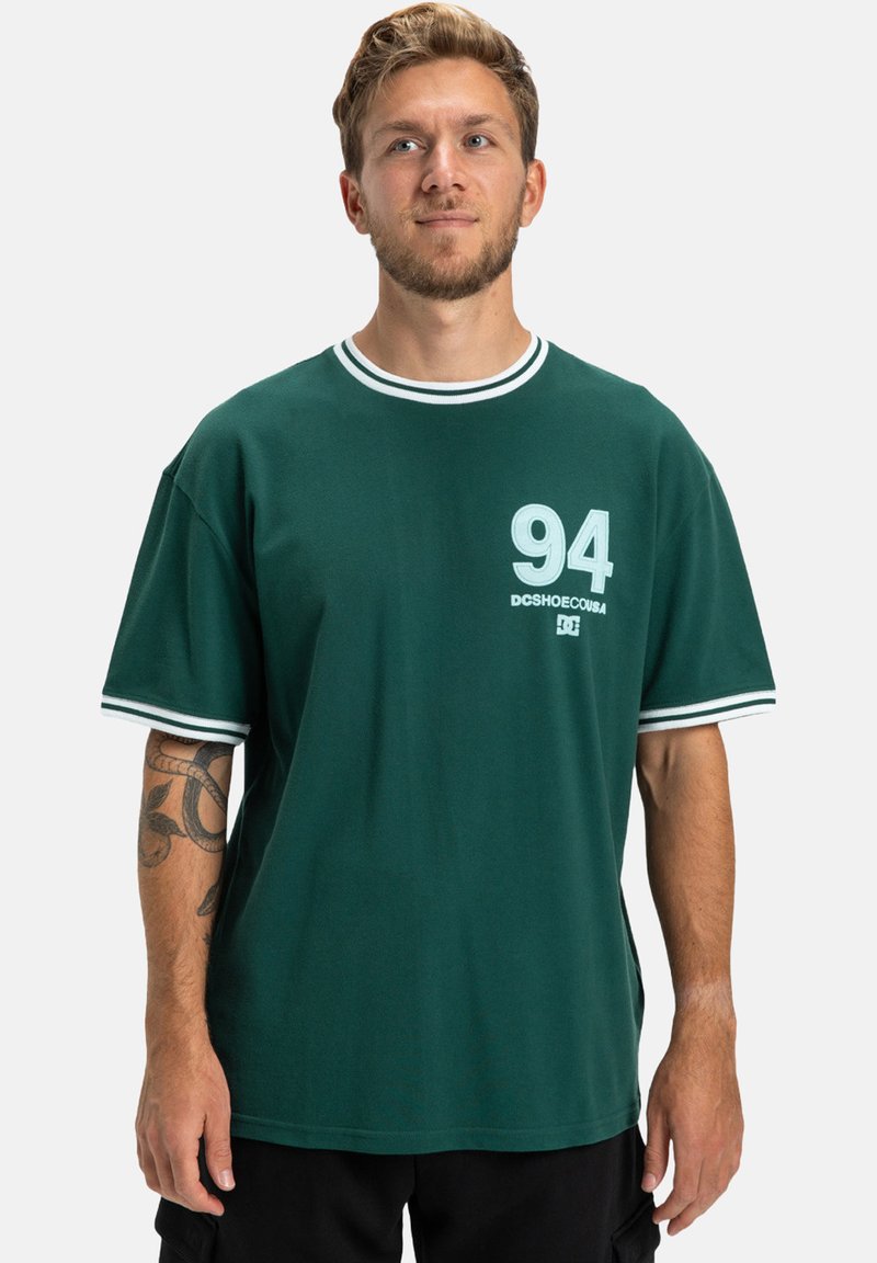 Mężczyzna z krótkimi włosami i brodą nosi ciemnozieloną koszulkę z białymi paskami przy kołnierzy i rękawach, z numerem 94 oraz tatuażem na lewej przedramieniu.