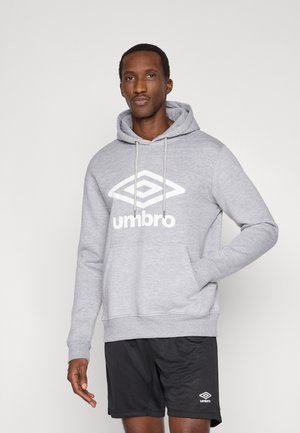 Mężczyzna ubrany w szarą bluzę z kapturem Umbro z logo oraz czarne spodenki Umbro, stoi na jednolitym jasnym tle z jedną ręką w kieszeni bluzy.