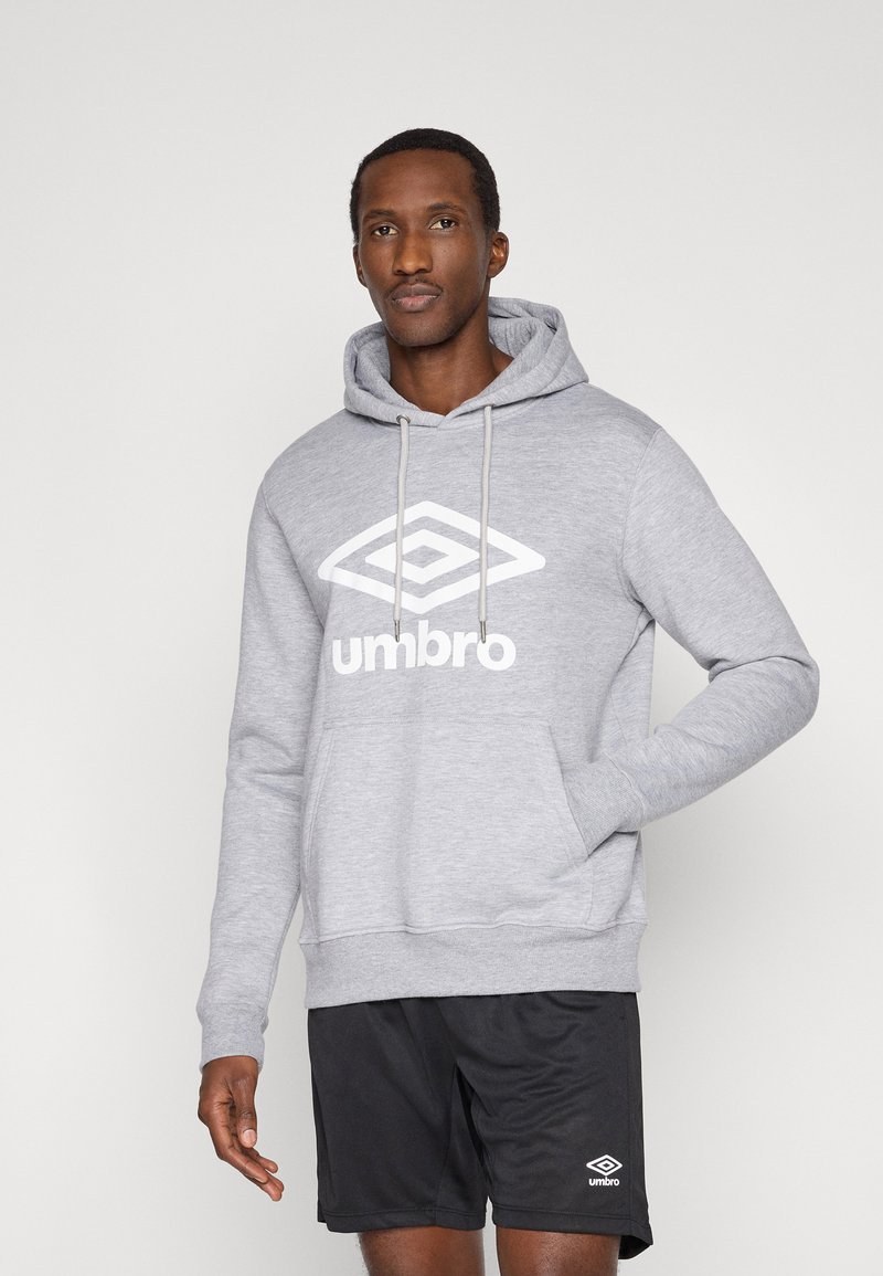 Homme portant un sweat à capuche Umbro gris avec logo et un short Umbro noir, debout devant un fond clair uni, une main dans la poche du sweat.