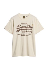 Cremefarvet bomulds T-shirt med stort vintage-stil brunt tekst og logo på forsiden. Korte ærmer, rund hals, afslappet pasform.