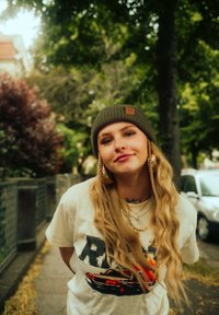 Braided hair, green knit beanie, cream t-shirt with graphic design, layered gold necklaces, hoop earrings; urban background with trees.