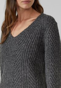 Grauer Strickpullover mit V-Ausschnitt, der ein strukturiertes Muster und einen subtilen Schimmer aufweist. Lange Ärmel und eine figurbetonte Silhouette.