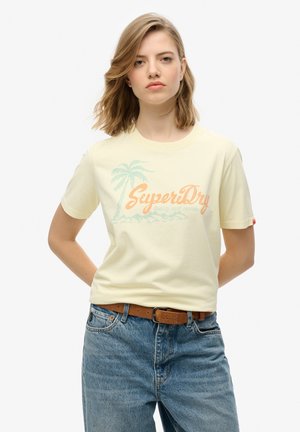 T-shirt à manches courtes jaune pâle avec un graphique de palmier bleu et le texte orange "SuperDry". Porté avec un jean bleu clair et une ceinture marron.