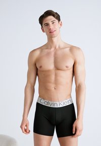 Fekete boxer alsónadrág szürke rugalmas derékszíjjal, amelyen a "Calvin Klein" logo látható. A modellen semleges kifejezés van, és izmos alkatú.