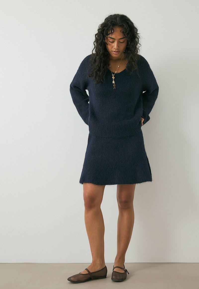 Vrouw met lang krullend haar, gekleed in een donkerblauwe gebreide trui en bijpassende rok, staand met de handen achter de rug en naar beneden kijkend.