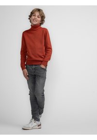 Maglione a dolcevita rosso, tessuto testurizzato, abbinato a jeans grigi aderenti e sneakers bianche con dettagli rossi, in piedi su uno sfondo uniforme.