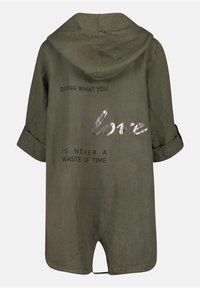 Oliveová zelená lněná kombinéza s kapucí, s textem v černé a stříbrné fólii: "DĚLAT TO, CO MÁTE RÁDI, NIKDY NENÍ ZTRÁTA ČASU." Krátké rukávy.