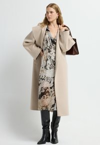 Beige uldblandingsfrakke med brede ærmer, båret over en mønstret mørk kjole. Sorte støvler til knæene og en lille brun håndtaske fuldender looket.