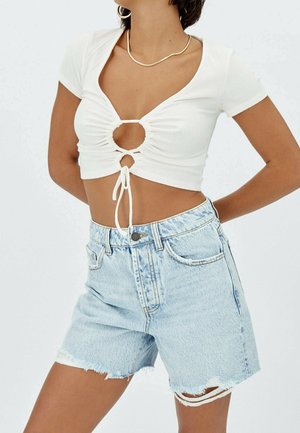 Top corto blanco de canalé con un diseño de ojal en la parte delantera y trasera, con lazos ajustables. Combinado con shorts de denim azul claro con dobladillos deshilachados.