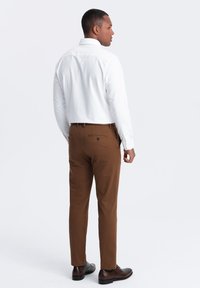 Weißes Hemd mit geradem Rücken, kombiniert mit braunen Hosen und dunklen Schuhen. Das Outfit zeichnet sich durch ein klares, modernes Design aus.