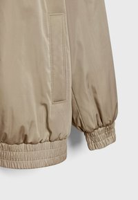 Beige bomber jacket with a smooth, glossy finish. Features elasticized cuffs and a side pocket. Simple, clean design without additional patterns.