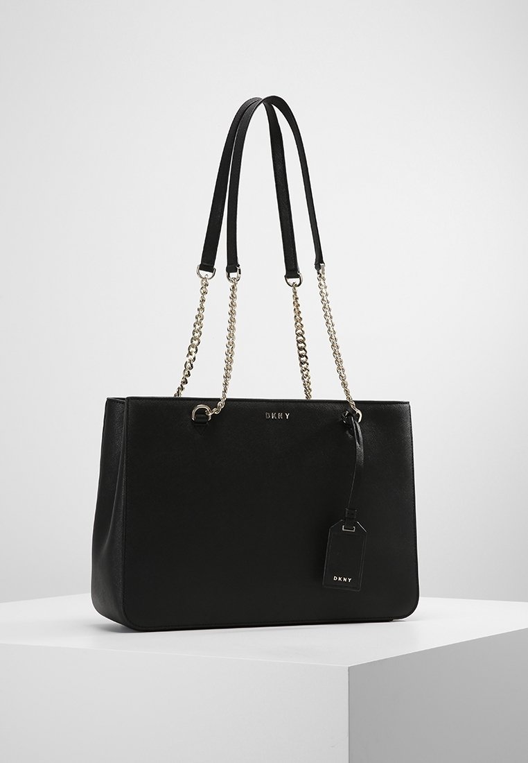 Sac fourre-tout en cuir noir avec doubles bretelles d'épaule, accents de chaîne argentée, détail du logo et une étiquette détachable. Texture lisse, design rectangulaire.