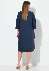 Donkerblauwe jurk met driekwart mouwen, een kraag en knoopdetails. De stof lijkt glad en heeft een knielange snit. Gemaakt met lichtgekleurde hakken.