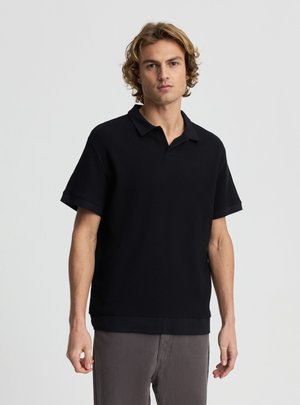 Giovane uomo con capelli biondi e ricci, indossa una polo nera a maniche corte e pantaloni grigi, in piedi davanti a uno sfondo chiaro e uniforme.