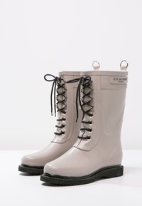 Bottes en caoutchouc grises avec un haut tige, des lacets noirs, des œillets en métal et des poignées en haut. Semelles noires robustes présentant une légère texture.