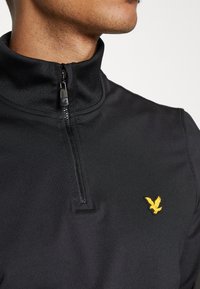 Zwart kwartel-rits pullover gemaakt van gladde stof, met een gele geborduurde logo van een adelaar op de linkerkant van de borst.