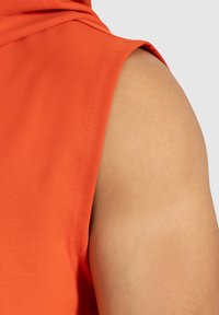 Camisetta arancione senza maniche con una texture liscia e aderente e cuciture a punto singolo, che mette in evidenza un primo piano della zona della spalla.