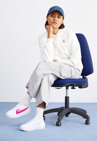 Bílé tenisky Nike s růžovým znakem, světle šedé kalhoty cargo a bílá mikina s logem. Modrá džínová čepice. Sedí na černé kancelářské židli.