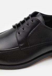 Scarpa in pelle nera con texture liscia, punta arrotondata e design con allacciatura a cinque occhielli. Presenta cuciture sottili e una suola a profilo basso.