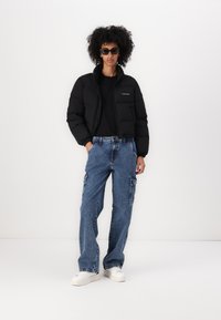 Zwarte pufferjack met ritssluiting, gecombineerd met baggy lichtblauwe denim cargobroek. Model draagt witte sneakers en zwarte zonnebril.