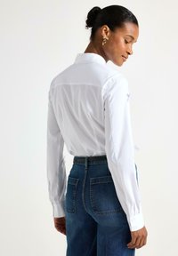 Camicia bianca con colletto e maniche lunghe con polsini abbottonati, abbinata a jeans in denim blu a vita alta. Legature note alle maniche.