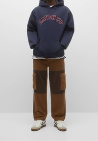 Person iført navy hoodie med teksten "CATCH UP", brune patchwork cargo bukser og hvide sneakers mod en ensfarvet baggrund.