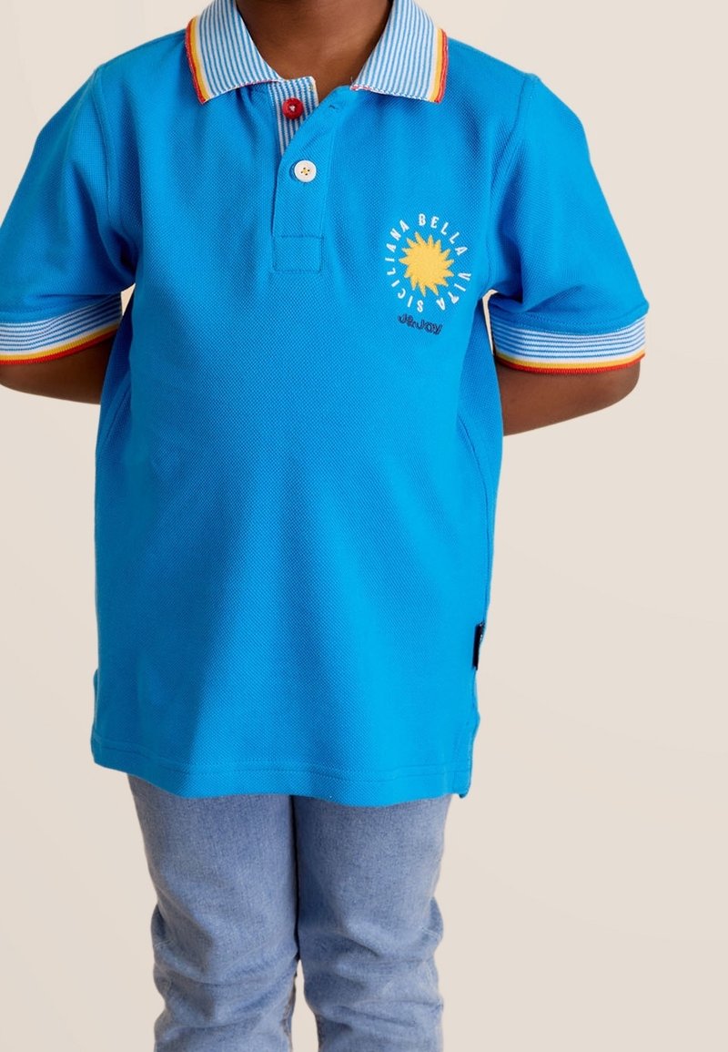 Enfant portant un polo bleu avec un col et des poignets rayés, un motif de soleil jaune sur la poitrine et un pantalon bleu clair, debout les mains derrière le dos.